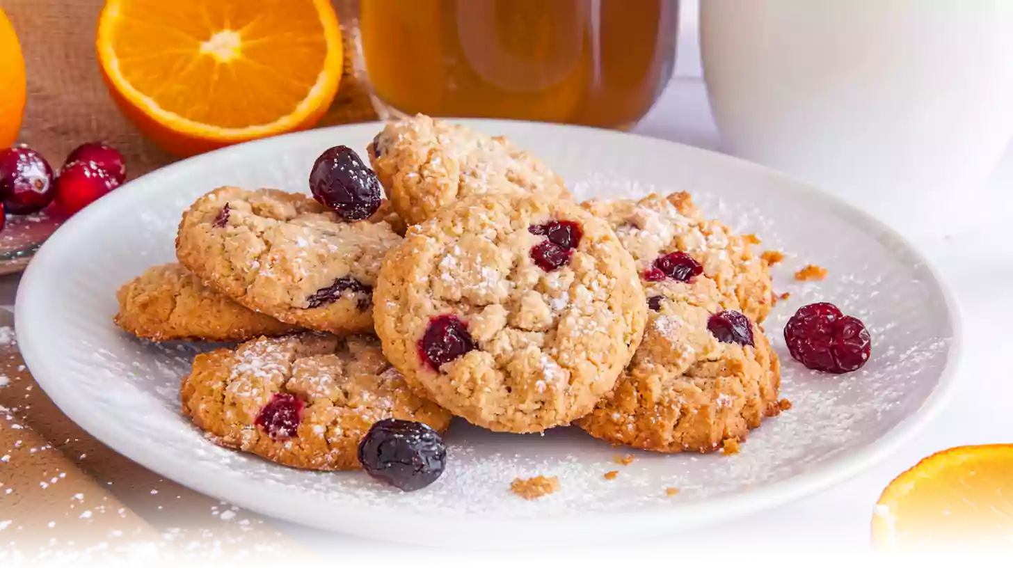 A plate of golden-brown orange and cranberry shortbread cookies dusted with powdered sugar, accompanied by fresh orange slices and a mug of tea.