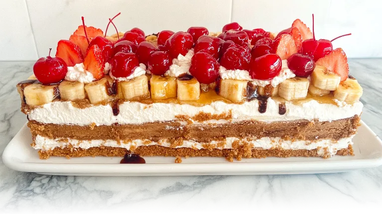 A classic banana split cake, beautifully layered with fruits and cream, garnished with cherries and chocolate drizzle.