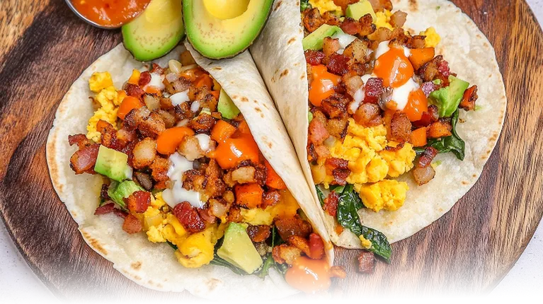 A perfectly sliced breakfast burrito showcasing layers of eggs, potatoes, bacon, cheese, and fresh veggies, served with avocado and salsa.