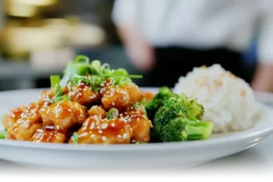 Glossy orange chicken garnished with sesame seeds and scallions on a modern plate, paired with rice and broccoli.