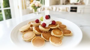 Golden mini pancakes stacked with fresh berries, whipped cream, and syrup in a modern kitchen setting.