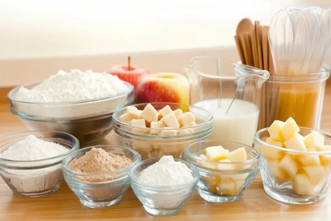 Ingredients for cake apple fritters, including flour, sugar, cinnamon, milk, eggs, and diced apples, arranged neatly in a modern kitchen.